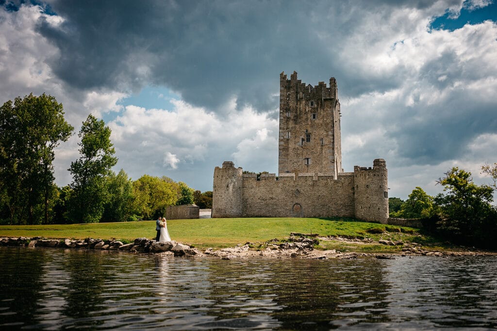 A newly wed couple in the their wedding clothes on the lakeside of Lough Leane with the lake in front of them and Ross Castle behind them. The photo was taken from a Gap of Dunloe Tours boat by Photographer Konrad Paprocki